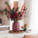 Het Zijden kunstbloemen boeket Cozy Winter in een mauve vaas, met roze bloemen, oranje bessen en dennentakken, siert een rond houten dienblad op een witte tafel voor een gezellige, moderne herfst/winterlook met houten stoelen en beige gordijnen.
