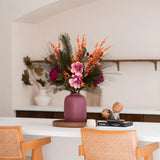 Een mauve vaas met het Zijden kunstbloemen boeket Cozy Winter - perfect voor herfst winterdecoratie - staat op een witte toonbank tussen houten stoelen, met boeken, artisjokken, planken en een witte pot met takken in de buurt.