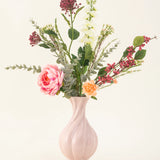 Het Zijden kunstbloemen boeket Pretty Petite, in een pastelroze vaas, bevat handgemaakte roze en oranje bloemen, witte bloemen, groene bladeren en rode bessen - perfect voor een duurzaam interieur tegen een effen lichte achtergrond.