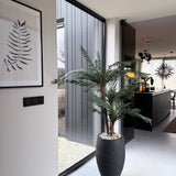 Een modern, minimalistisch interieur met een Kunstpalm Cycas Classic 135 cm in een stijlvolle sierpot bij een raam van vloer tot plafond, een varen-kunstwerk aan de muur en een strakke zwarte keuken met hanglampen op de achtergrond.