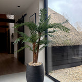 De Kunstpalm Cycas Classic 135 cm in een strakke sierpot staat bij een groot raam met daglicht. Buiten zijn een rieten dak en grind te zien. Het interieur is modern en minimalistisch.