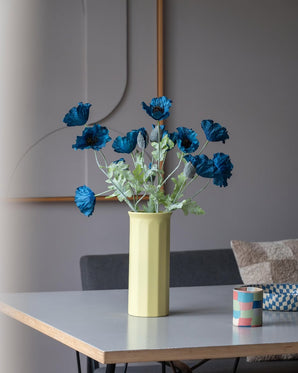 Een Kunstbloem Poppy van 73 cm in donkerblauw staat in een gele vaas op een tafel, vergezeld van een klein, kleurrijk gestreept kopje, in een moderne, zacht verlichte kamer met een minimalistische inrichting en neutrale tinten.