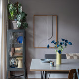 De moderne eethoek is voorzien van een grijze glazen vitrinekast waarin serviesgoed en planten zijn uitgestald, minimalistische wanddecoraties en een tafel waarop de Kunstbloem Poppy 73 cm donkerblauw in een gele vaas staat. De ruimte wordt gecompleteerd door een eclectische mix van stoelen, allemaal tegen een achtergrond van grijze muren en een houten vloer.