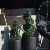 Twee Kunst Buxus Spiraal Romeinse stijl UV 90cm vormsnoeiplanten met spiraalvormige bladeren staan op een betonnen patio naast een zwarte hangstoel en een moderne buitenbank op de achtergrond.