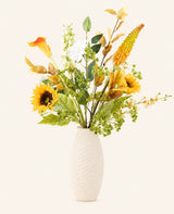 Het Zijden kunstbloemen boeket Amber Light, gerangschikt in een gestructureerde witte vaas, heeft zonnebloemen, witte en gele bloemen, groene bladeren en gebladerte tegen een lichte achtergrond.