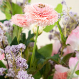 Een close-up van het Zijden kunstbloemen boeket Belle Michelle, met een roze ranonkel, omringd door lichtpaarse en roze bloemen met groene bladeren op de achtergrond.