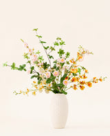 In een vaas met witte structuur staat het Zijden kunstbloemen boeket Blossom Harmony met roze, witte en gele kunstbloemen en groene bladeren tegen een effen lichte achtergrond.