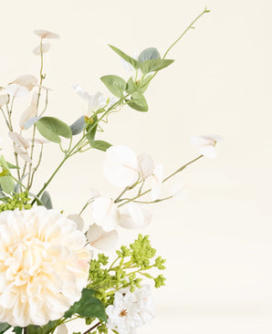 Een close-up van het Zijden kunstbloemen boeket Claire de Lune, met witte en crèmekleurige bloemen, groene bladeren en delicaat wit gebladerte op een lichte, neutrale achtergrond.