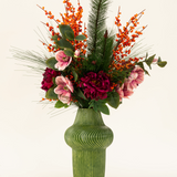 Het Zijden kunstbloemen boeket Cozy Winter presenteert een groene ribbelvaas met donkerrode en roze bloemen, oranje bessen, dennentakken en groene bladeren - perfect als herfst-winter decoratie tegen een lichte achtergrond.