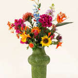 Een groene vaas met structuur toont het Zijden kunstbloemen boeket Colourful Spirit, met zonnebloemen, roze pioenrozen, oranje en paarse bloemen en weelderig groen tegen een effen lichte achtergrond.