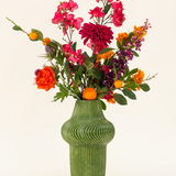 Het Zijden kunstbloemen boeket Fiesta Sunset is gerangschikt in een groene vaas met textuur en toont roze, oranje en paarse bloemen met groene bladeren en kleine oranje vruchten tegen een lichte achtergrond.