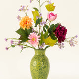 Een groen gespikkelde vaas toont het Zijden kunstbloemen boeket Love in Bloom, met roze, oranje en donkerrode bloemen met paars en groen gebladerte tegen een lichte achtergrond.