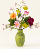 Een groen gespikkelde vaas toont het Zijden kunstbloemen boeket Love in Bloom, met roze, oranje en donkerrode bloemen met paars en groen gebladerte tegen een lichte achtergrond.