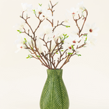 Een groene vaas toont het Zijden kunstbloemen boeket Magnolia wit, met verschillende witte magnoliatakken met kleine groene blaadjes, tegen een effen lichte achtergrond.