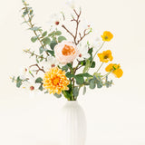 Het Zijden kunstbloemen boeket Fields of Joy heeft gele, witte en perzikkleurige bloemen met groene eucalyptus in een vaas met witte textuur, prachtig gerangschikt tegen een lichte achtergrond.