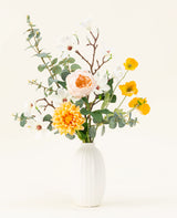 Het Zijden kunstbloemen boeket Fields of Joy heeft gele, witte en perzikkleurige bloemen met groene eucalyptus in een vaas met witte textuur, prachtig gerangschikt tegen een lichte achtergrond.