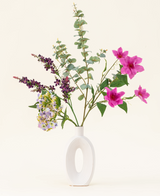 Het Zijden kunstbloemen boeket Lavender Skies, gerangschikt in een moderne witte vaas, bevat roze en paarse kunstbloemen, gele accenten en weelderig groen gebladerte - prachtig tegen een effen lichte achtergrond.