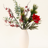 Het Zijden kunstbloemen boeket Winterberry Bliss heeft rode bessen, dennentakken, eucalyptus, rode amaryllis en dennenappels in een witte vaas met textuur - ideaal voor kerstdecoratie of om te laten zien tegen een lichte achtergrond.