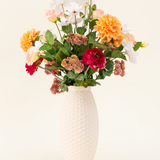 Het Zijden kunstbloemen boeket The Wild Bunch, met rode rozen, Real Touch Dahlia, roze bloemen en groene bladeren in een vaas met witte structuur, wordt getoond tegen een lichte achtergrond.