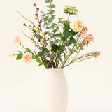 Een vaas met witte structuur toont het Zijden kunstbloemen boeket Blush Blossom, met echte bloemen - lichtroze rozen, gele bloemen en groen gebladerte - tegen een effen lichte achtergrond.