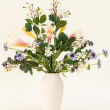In een witte vaas staat het Zijden kunstbloemen boeket Blush & Bloom - met roze lelies, witte en paarse bloemen en weelderige groene bladeren - tegen een lichte achtergrond.