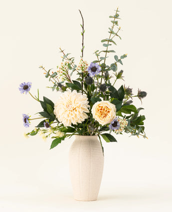 Een witte vaas van keramiek bevat het Zijden kunstbloemen boeket Petal Poetry, met crèmekleurige dahlia's en rozen, lichtpaarse bloemen, groene distels en allerlei groen tegen een effen lichte achtergrond.