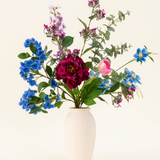 Een vaas met witte structuur toont het Zijden kunstbloemen boeket Painted Petals, met levendige blauwe, paarse, roze en rode zijden bloemen met groene bladeren en eucalyptustakken tegen een effen lichte achtergrond.