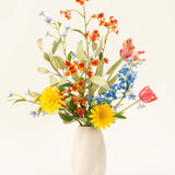 Het Zijden kunstbloemen boeket Lemonade Garden heeft echt getinte zonnebloemen, roze tulpen, blauwe riddersporen, oranje bloesems en groen gebladerte in een beige vaas tegen een effen lichte achtergrond.