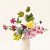 Het Zijden kunstbloemen boeket Aurora Garden heeft echte aanraakbloemen in roze, geel, blauw en paars met groene bladeren, gerangschikt in een witte keramieken vaas tegen een effen lichte achtergrond.
