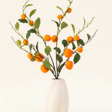 Het Zijden kunstbloemen boeket Mandarijn heeft een vaas met witte textuur en takken van groene bladeren en levendige oranje kunstmandarijnen, die een levendige toets geven tegen een effen lichte achtergrond.