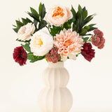 Een crèmekleurige vaas van keramiek met structuur toont het Zijden kunstbloemen boeket Jewel Bloom met roze, witte en donkerrode Real Touch bloemen en groene bladeren tegen een effen lichte achtergrond.