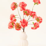 Het Zijden kunstbloemen boeket Kersenbloesem Koraal heeft levendige roze en koraal kunstmatige kersenbloesems met groene bladeren, elegant gerangschikt in een wit gebeeldhouwde vaas tegen een lichte achtergrond.