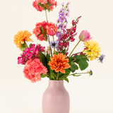 Het Zijden kunstbloemen boeket Colourburst Blossom heeft een pastelroze vaas gevuld met oranje dahlia's, roze anjers, gele dahlia's, paarse riddersporen en rode tulpen-ideale kunstboeket decoratie voor elke kamer.