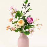 Het Zijden kunstbloemen boeket Touch of Cloudlight, gepresenteerd in een roze keramieken vaas, bevat roze pioenrozen, gele rozen, kleine bloesems, groen en takken met donkere bessen tegen een lichte achtergrond.