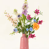 Het Zijden kunstbloemen boeket Bloom & Glow heeft roze, paarse, blauwe, oranje, witte en gele bloemen met groene bladeren in een roze vaas van keramiek tegen een lichte achtergrond.