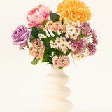 Een witte vaas van gegolfd keramiek toont het Zijden kunstbloemen boeket Peach Delight, met een gele dahlia, roze pioenroos, paarse roos en verschillende kleine witte en roze bloemen tegen een eenvoudige lichte achtergrond.