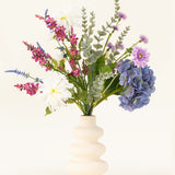 Een crèmekleurige golvende vaas bevat het Zijden kunstbloemen boeket Lavender Kiss, met witte dahlia's, lila kunstbloemen, paarse hortensia's, roze en paarse bloemen en groen gebladerte tegen een effen lichte achtergrond.