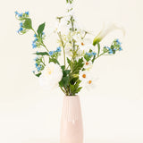 De lichtroze vaas bevat het Zijden kunstbloemen boeket Whispers of White, met witte bloemen, blauwe bloesems en groen gebladerte tegen een effen, lichte achtergrond.