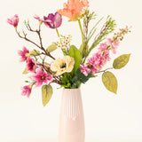 Het Zijden kunstbloemen boeket Spring Awakening, met roze magnolia's, lichtgele klaprozen en oranje en paarse bloemen in een hoge lichtroze keramieken vaas, benadrukt het Nederlands vakmanschap tegen een effen witte achtergrond.