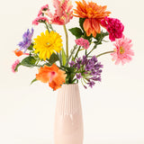 Een lichtroze vaas van keramiek bevat het Zijden kunstbloemen boeket Candy Petals met levendige oranje, gele, roze en paarse zijden bloemen tegen een effen gebroken witte achtergrond.