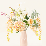 Een pastelroze vaas van keramiek toont het Zijden kunstbloemen boeket Blushing Beauty, met zachtgekleurde hortensia's, rozen en divers groen, prachtig gerangschikt tegen een bleke neutrale achtergrond.