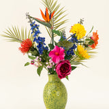 Een groene, gespikkelde vaas toont het Zijden kunstbloemen boeket Hibiscus Fiesta - met roze, rode en gele bloemen, blauwe riddersporen, oranje paradijsvogels en groene palmbladeren - tegen een effen lichte achtergrond.