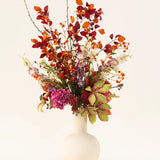 Het Zijden kunstbloemen boeket Golden Autumn heeft een kunstzinnige mix van herfstbladeren en rode, oranje en paarse bloemen in een witte keramieken vaas, tegen een lichte achtergrond.