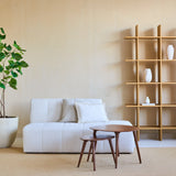 Minimalistische woonkamer met een witte bank, twee geneste houten salontafels, beige muren, een open houten plank met witte vazen en een Kunstplant Ficus 180 cm die groen toevoegt. Een beige vloerkleed bedekt de vloer.