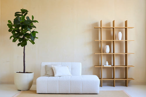 Minimalistische woonkamer met een witte bank, twee kussens en een Kunstplant Ficus Tabaksplant Deluxe 180 cm naast een open houten plank met witte vazen, alles tegen een beige muur en op een licht vloerkleed.