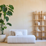 Minimalistische woonkamer met een Kunstplant Monstera 240 cm in een pot, een moderne witte bank met kussen en een houten open kast met witte vazen tegen een beige muur. Natuurlijke lichtinval van rechts.
