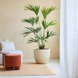 De Kunstplant Kentia Palm 180 cm staat op een bruin vloerkleed naast een witte bank met kussens en een kleine ronde oranje voetenbank in een lichte, minimalistische kamer met transparante witte gordijnen.