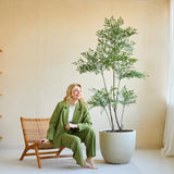 Een vrouw in een groen pak glimlacht terwijl ze op een houten stoel naast de Kunstplant Ruscus 180 cm zit in een lichte, moderne kamer met beige muren, grote ramen en een minimalistische inrichting.