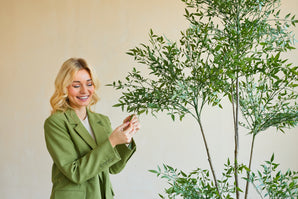 Een glimlachende vrouw in een groene blazer staat binnen en raakt voorzichtig de bladeren aan van een Kunstplant Ruscus 150 cm met dunne takken en weelderig groen gebladerte, dat afsteekt tegen een effen, lichtgekleurde achtergrond.