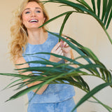 Een glimlachende vrouw met lang blond haar, in een lichtblauwe mouwloze top en bijpassende broek, staat binnen achter de Kunstplant Kentia Palm 180 cm en raakt speels een van de groene bladeren aan.